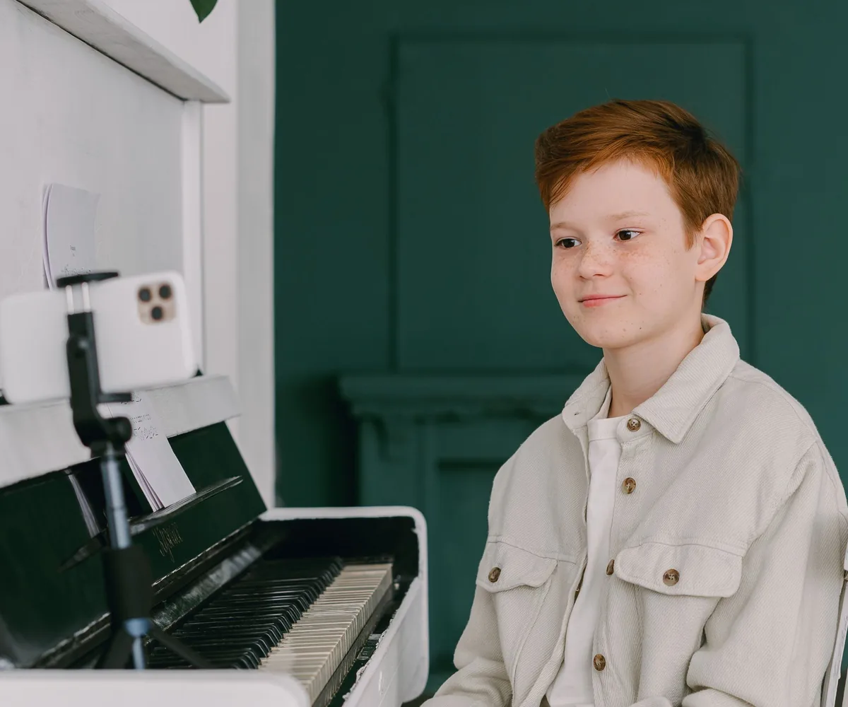 A boy at the piano smiling at his teacher on a phone screen during an online lesson